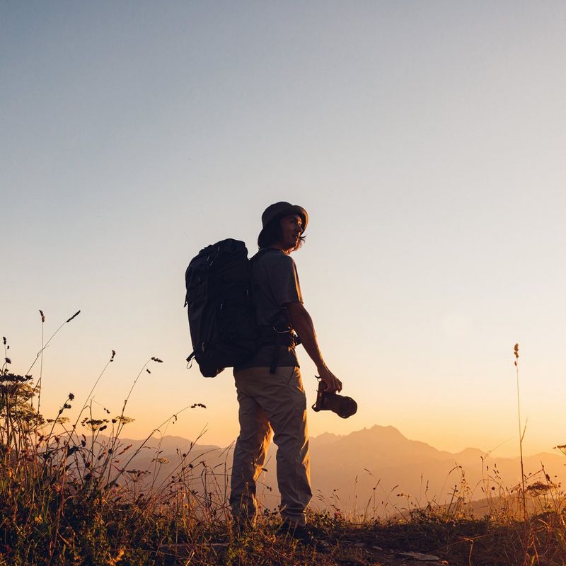 A man silhouetted against a sunset, wearing a backpack and holding a camera, in a natural outdoor setting.