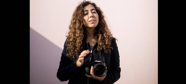 Photographer Tasneem Alsultan holds a Canon DSLR camera and lens in front of a pink wall.