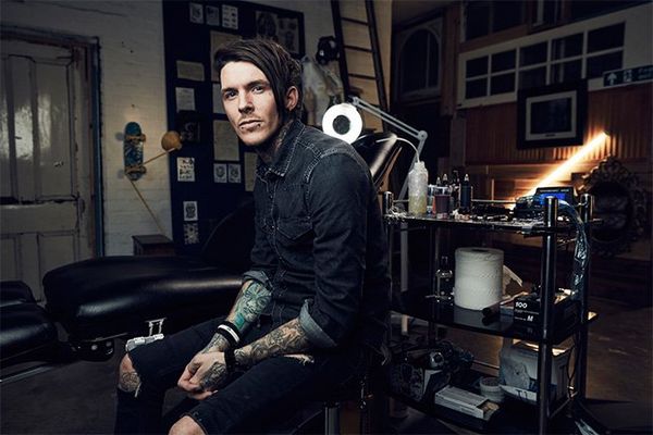 A tattooed man wearing denims sits in front of a trolley holding tattooist’s equipment