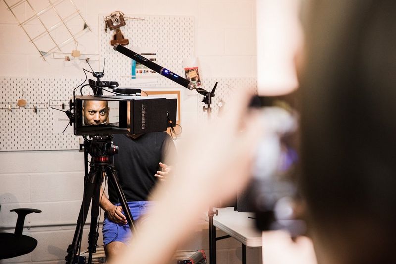 A man being photographed sat behind a video camera.