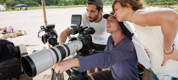 Two men and a woman look at the back of a Canon video camera with a long lens.