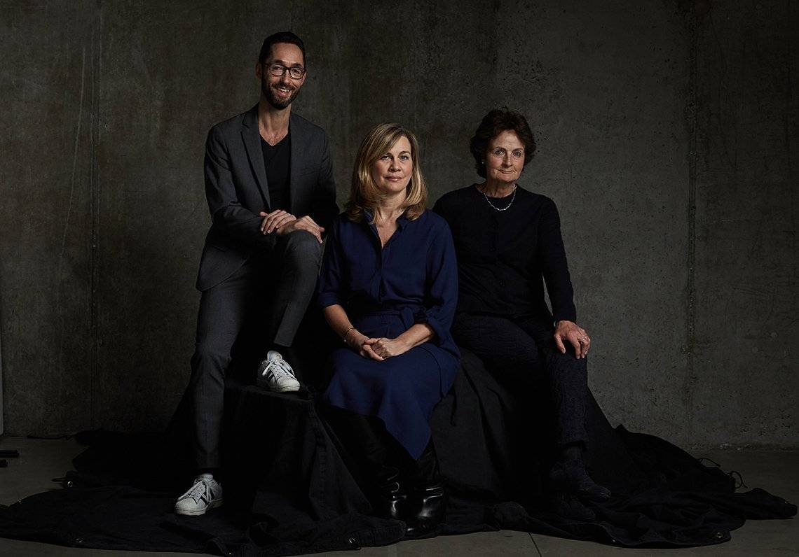 Thomas Borberg, Magdalena Herrera and Helen Gilks, all in smart clothing, sit against a grey backdrop on a black fabric-covered seat.