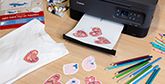 A Canon printer prints a sheet of paper with three heart designs on it. Around it are coloured pencils, a pack of transfer paper and a white t-shirt with a heart transfer on it.