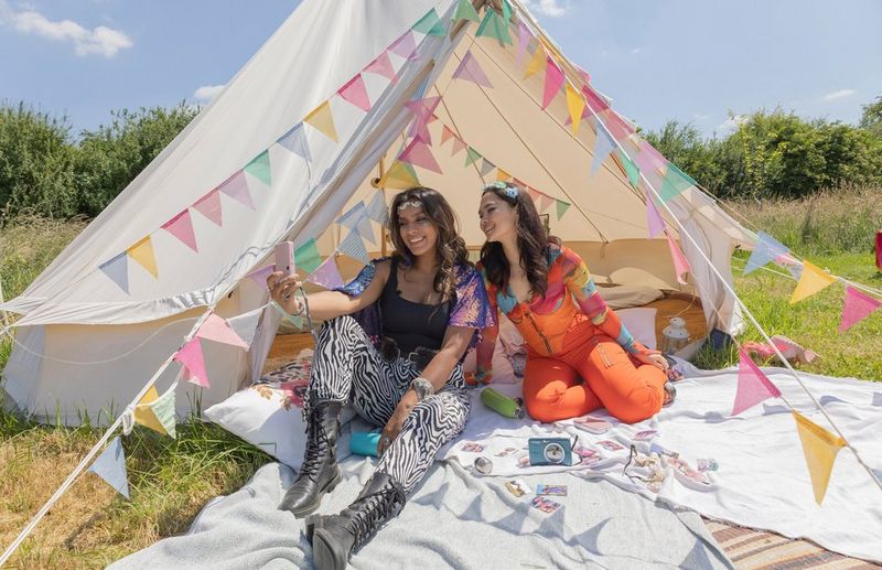 Two women in flamboyant clothing take a selfie with an instant camera while sitting at the entrance to a tent festooned with colourful bunting.