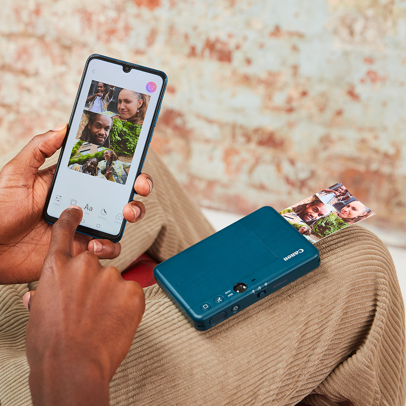 A man holding a phone with 4 pictures in a layout on it. He has Canon Zoemini S2 on his lap printing the image.