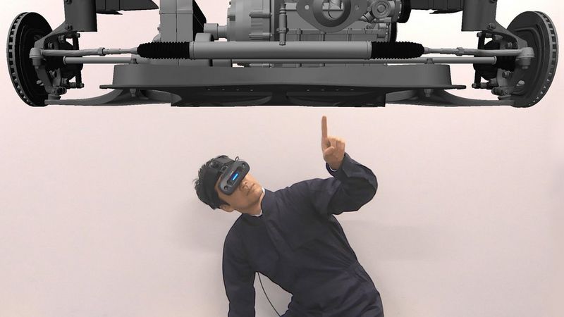 A man dressed in black is wearing a black Canon MREAL X1 mixed reality headset that covers his eyes. It has a neon blue stripe in the space between where his eyes should be. He crouches beneath a virtual representation of the chassis of a vehicle and points a finger to a spot just off the centre.