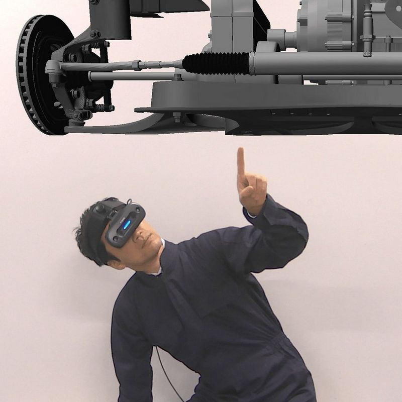 A man dressed in black is wearing a black Canon MREAL X1 mixed reality headset that covers his eyes. It has a neon blue stripe in the space between where his eyes should be. He crouches beneath a virtual representation of the chassis of a vehicle and points a finger to a spot just off the centre.