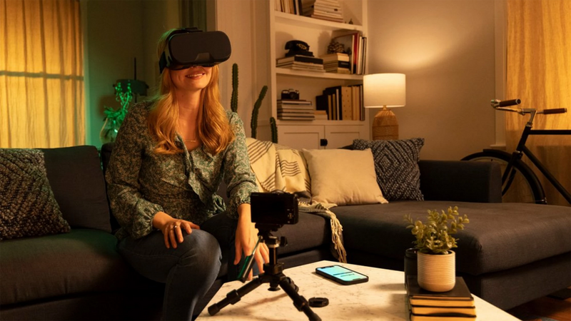A woman with long blonde hair sits on a couch. She wears a patterned green blouse and grey trousers, and a VR headset. In front of her is a coffee table, upon which is a camera on a tripod, a smartphone, some books and a small plant. Behind her is a white bookcase, two curtained windows and a bicycle. She’s holing the camera with a single hand as it rests on an orange tarpaulin. Besides the tarpaulin are ropes and beyond them is the sea. Together it appears that the photographer is on a boat at sea."