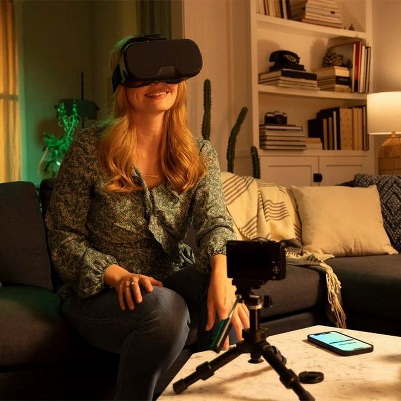 A woman with long blonde hair sits on a couch. She wears a patterned green blouse and grey trousers, and a VR headset. In front of her is a coffee table, upon which is a camera on a tripod, a smartphone, some books and a small plant. Behind her is a white bookcase, two curtained windows and a bicycle.