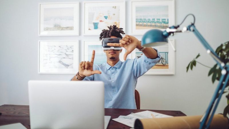 A man in a blue t-shirt is wearing a VR headset while sitting at a desk, a laptop opened in front of him. He makes a framing gesture with his hands. 