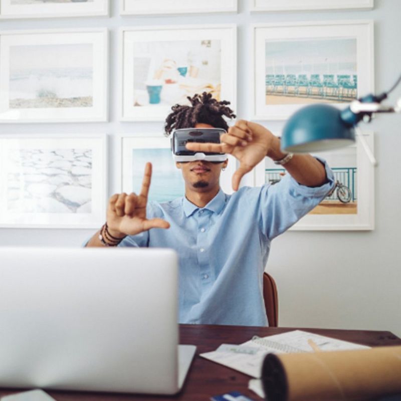 A man in a blue t-shirt is wearing a VR headset while sitting at a desk, a laptop opened in front of him. He makes a framing gesture with his hands.