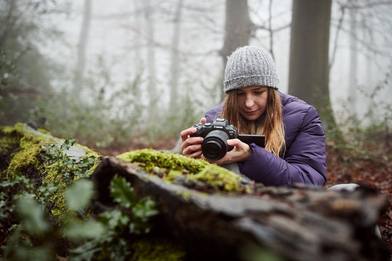 A woman in a warm hat and coat looking at the LCD screen of her Canon camera while crouching down to photograph a moss-covered rock.