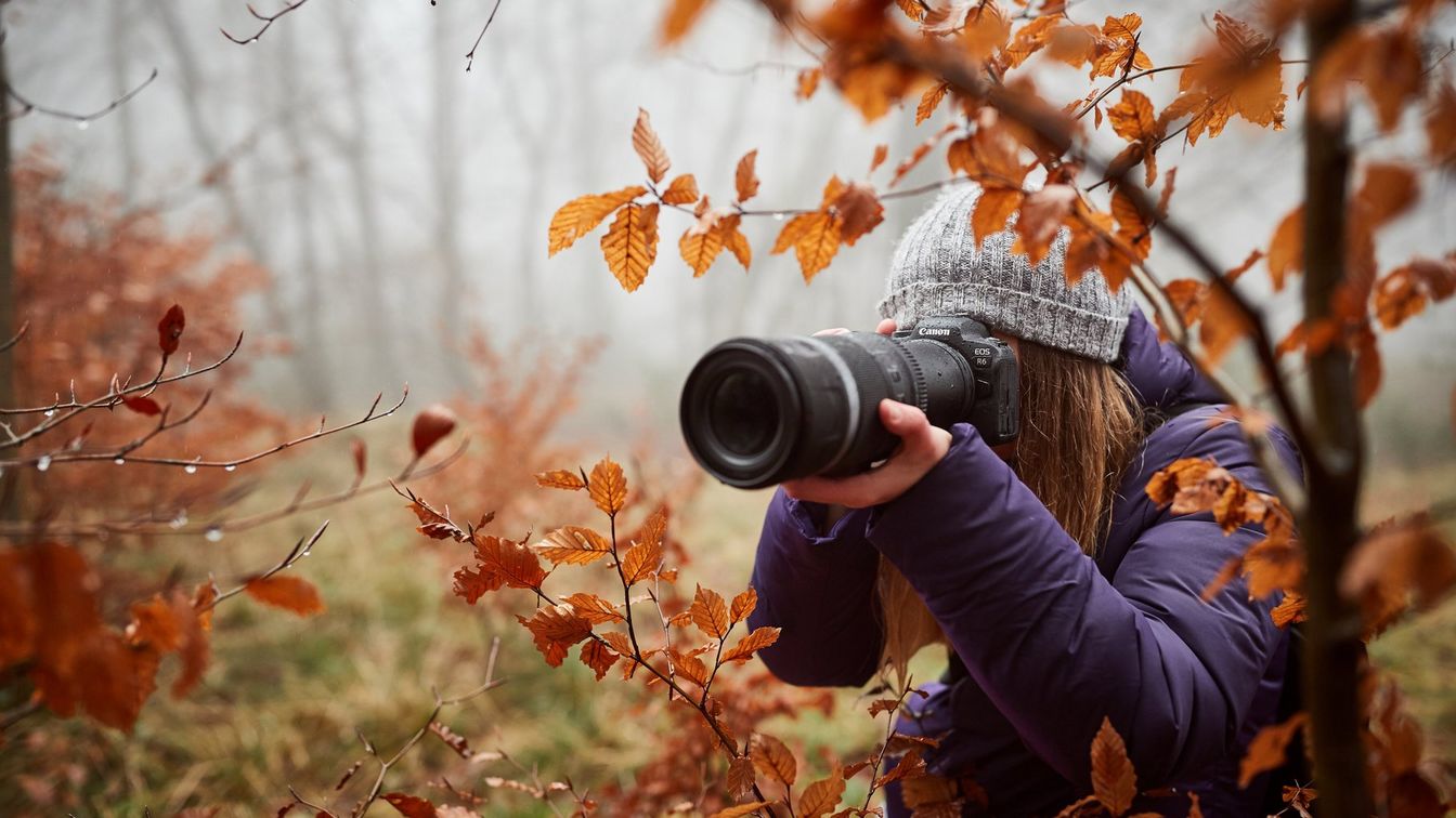 امرأة شابة ترتدي قبعة شتوية وهي تصور أوراق الخريف بكاميرا وعدسة كبيرة من Canon.