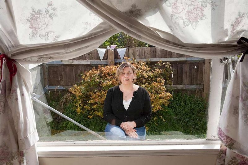 Elisabeth Blanchet is captured through the windowpane of a house, curtains framing the shot.