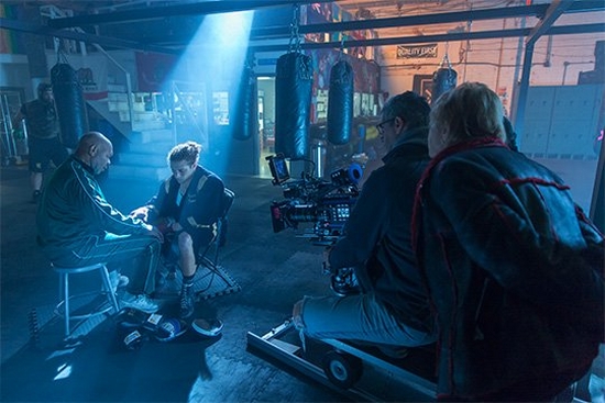 A boxer and trainer are lit from above by a shaft of light as they sit in a dim gym being filmed by a camera crew using a Canon EOS C700 FF.