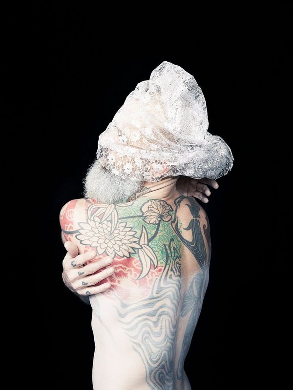 Matthew, from Felicity McCabe’s series Two Spirit. The bald, heavily tattooed man with a striking grey beard poses and wraps his arms around himself in the studio wearing a lace veil over his eyes.