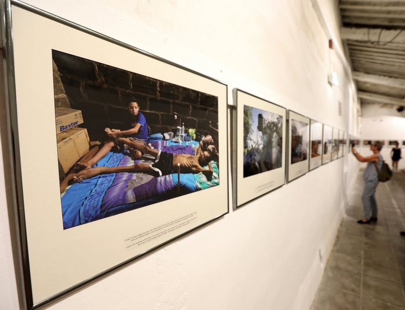 A close-up shot of a wall of framed photos, with a viewer taking a photograph of a picture in the distance.