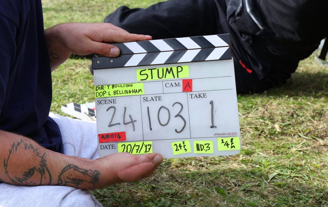 A close-up of a man holding a clapboard for Cam A with Scene 24 Slate 103 Take 1 written on it. He sits on grass and another person can be seen sitting on the grass behind him.
