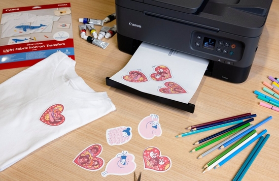 A Canon printer prints a sheet of paper with three heart designs on it. Around it are coloured pencils, a pack of transfer paper and a white t-shirt with a heart transfer on it.
