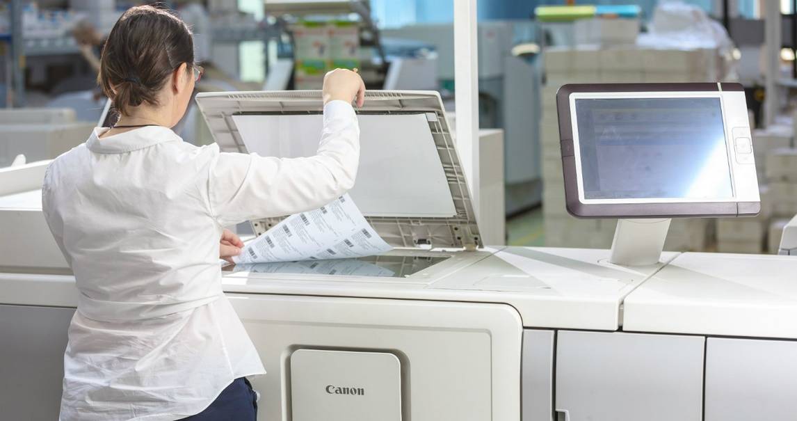 Woman with short dark hair wearing navy trousers and a long-sleeved white shirt uses a scanner on top of a Canon commercial digital printing press.