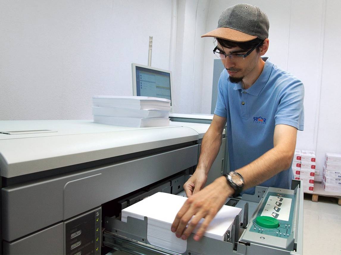 Man in sky blue polo shirt and a baseball cap loads A4 paper into the open drawer of a large Canon digital printing press.