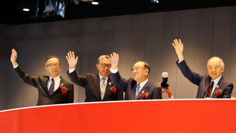 Four Japanese men in suits with red floral buttonholes stand behind a red balcony with their arms held up in a wave.