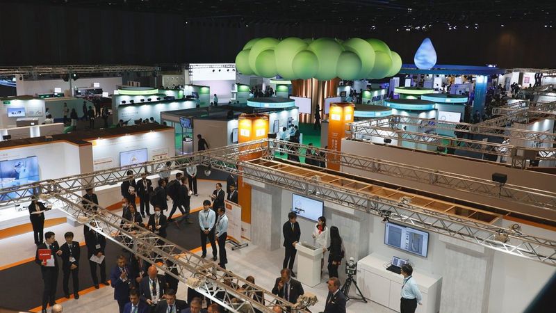 An aerial view of the Canon EXPO at Pacifico Yokohama North exhibition centre. Visitors standing at booths of technology surround a central brightly lit ‘tree’ formed of green cartoon-like bubbles atop a bronze trunk.