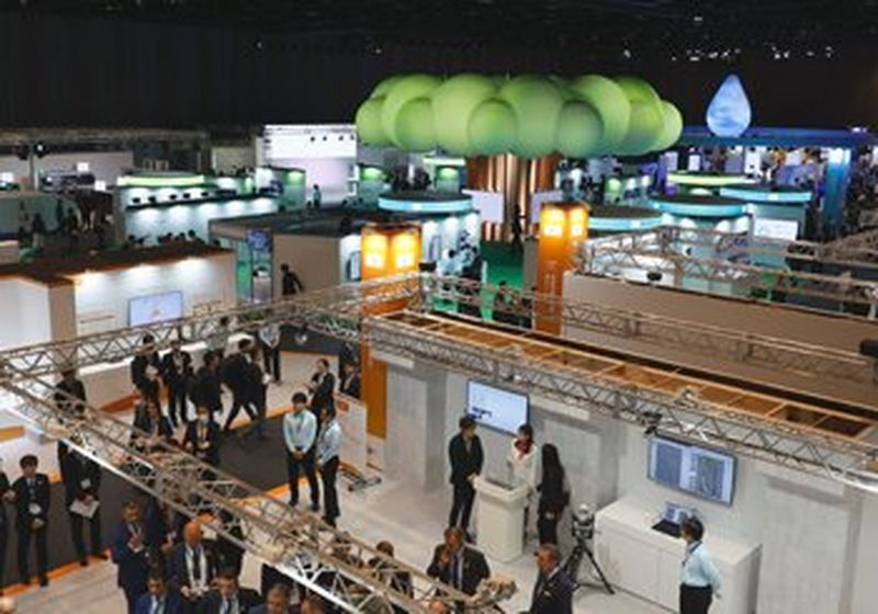 An aerial view of the Canon EXPO at Pacifico Yokohama North exhibition centre. Visitors standing at booths of technology surround a central brightly lit tree formed of green cartoon-like bubbles atop a bronze trunk.