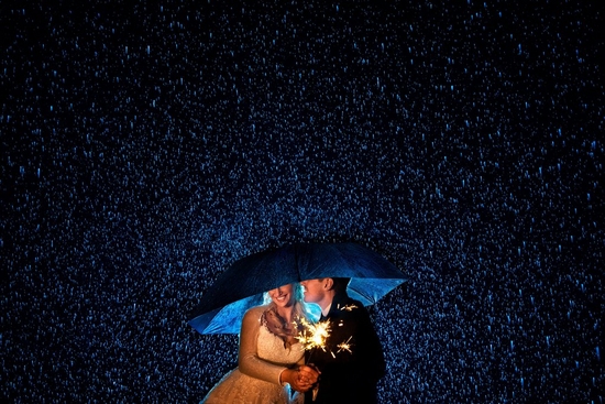 Newlyweds hold a sparkler while standing under an umbrella in the rain. 