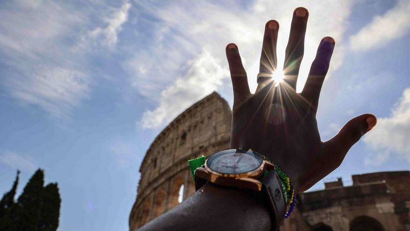 A hand reaches towards the sky above the Coliseum. A chunky analogue watch and beaded bracelets are visible on the person's wrist. There is a wedding band on their ring finger. A sun flare bursts from between the ring and middle fingers.