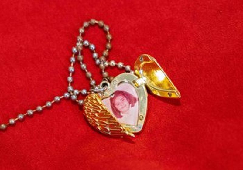 A gold, heart-shaped locket lies on a background of a blood red fabric. The front opening of the locket is two wings meeting and one wing is open, revealing an old, fading photograph of a little girl.