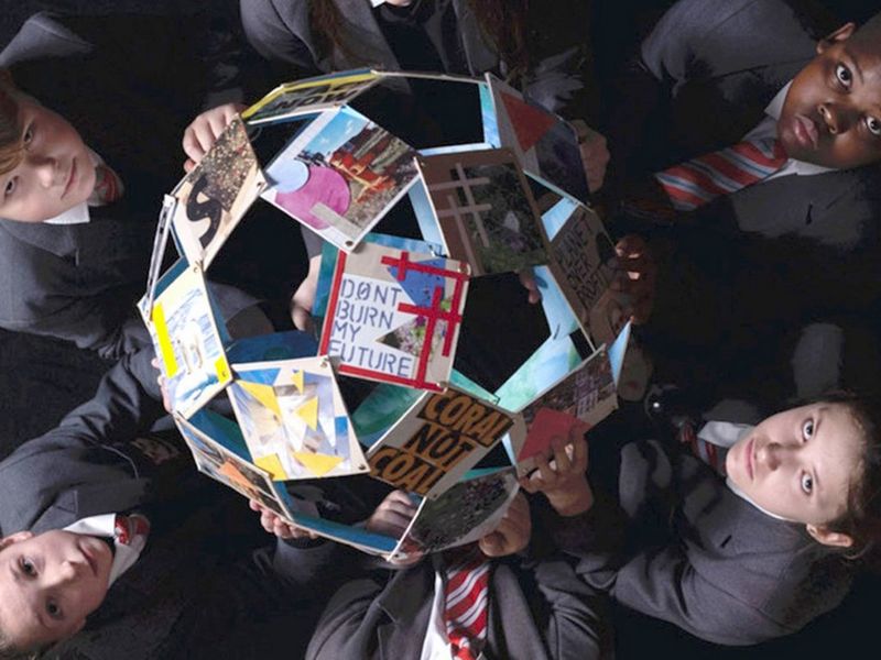 Taken from above, multiple children, on the left and right of the photo, hold a globe made of photographs and raise their solemn faces to the camera. The clearest photo, closest to the camera displays the words ‘don’t burn my future’.