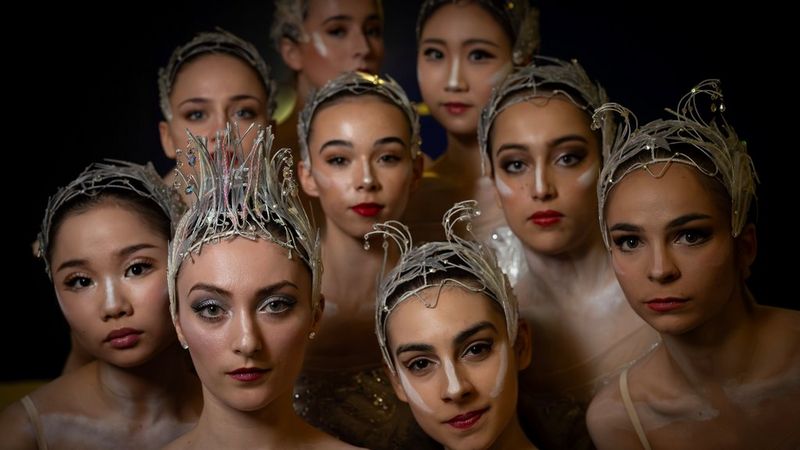 A group of ballet dancers in ornate headpieces and stage makeup pose closely together against a dark background.