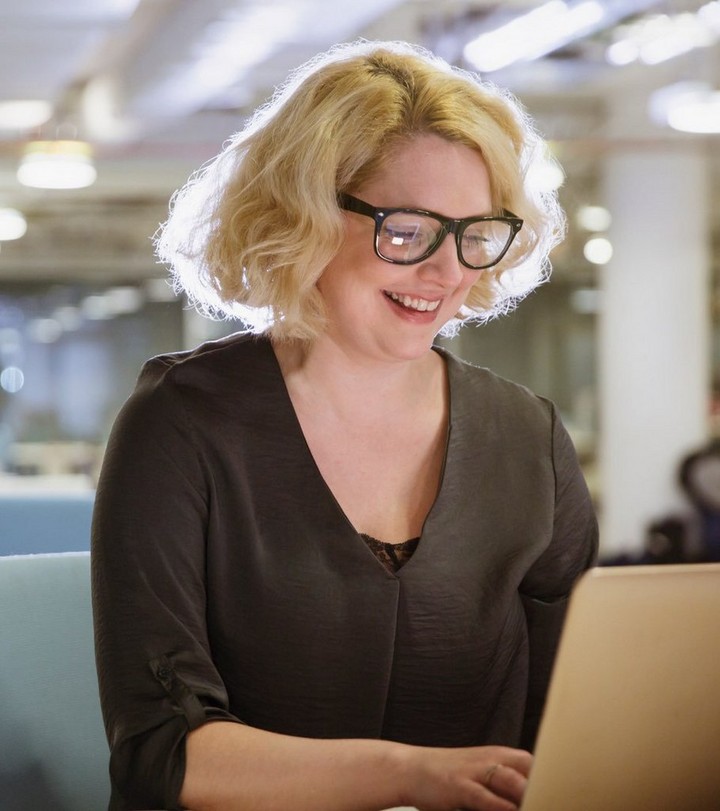 Blonde-haired woman in dark-rimmed glasses and khaki blouse smiles broadly as she looks at something on her laptop.