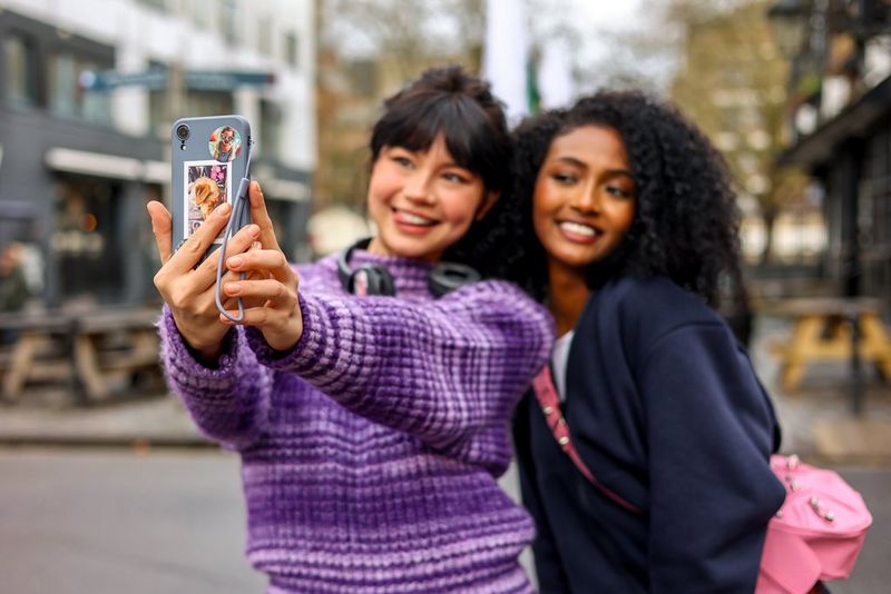 A woman in a purple jumper holds a smartphone to take a selfie while a woman in a dark blue jumper smiles next to her.