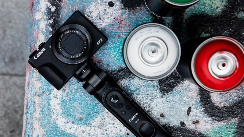 Canon PowerShot G7 X Mark III, kurai piestiprināts rokturis un kura novietota pie sienas blakus krāsas aerosola bundžām.