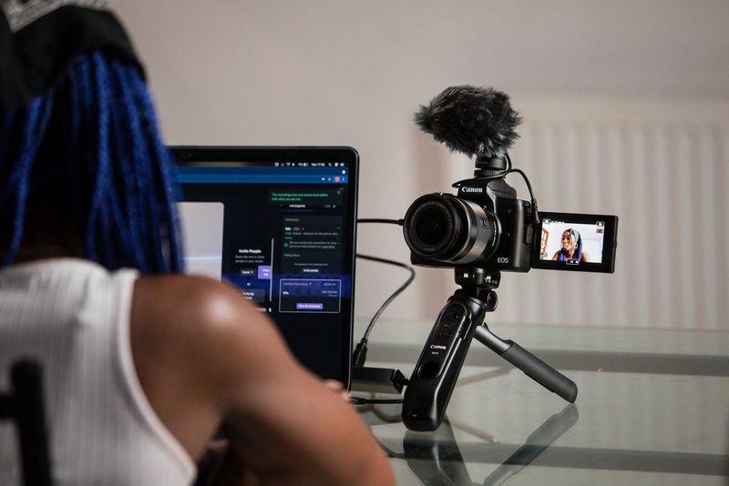 امرأة ذات شعر أزرق تجلس أمام كمبيوتر محمول بينما تلتقط كاميرا Canon مثبتة على حامل ثلاثي القوائم صورًا لها.