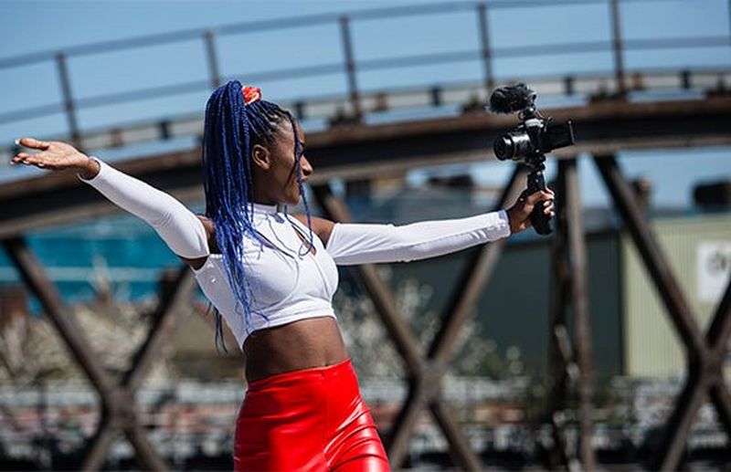 A young woman with blue braids filming herself with a camera on a handheld grip in front of a railway line.