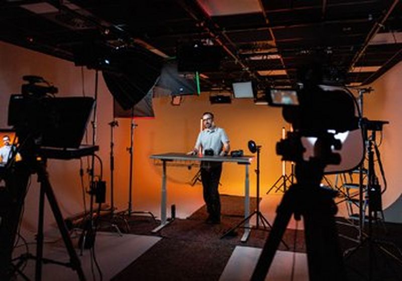 A bearded man stands behind a table in a film studio. There are lights and cameras trained on him.