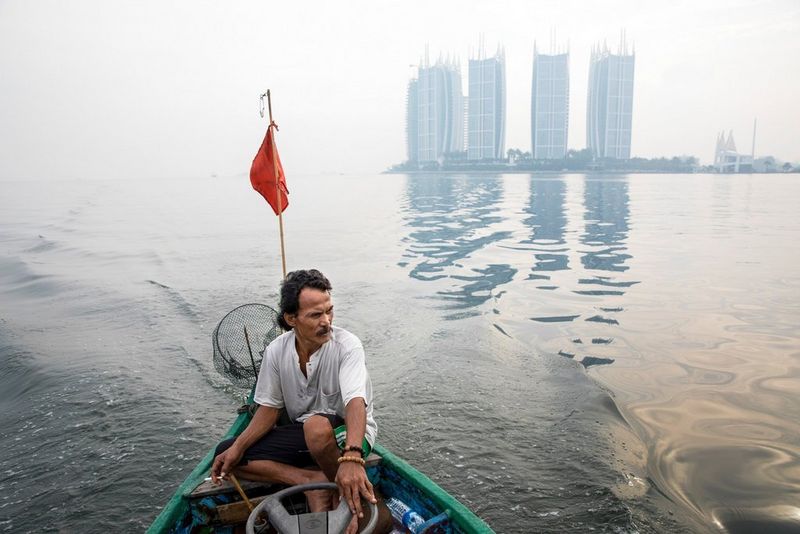 A man steers a small boat across a bay with tall skyscrapers in the background. Taken by Cynthia Boll. 