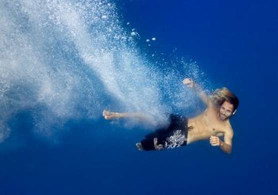A man in black swimming shorts, shown as he enters the water. He has clearly jumped or fallen in with some force, as the disturbance of white water can be seen behind him from his entry on the left. On the right, the water is clear, calm and deep blue.