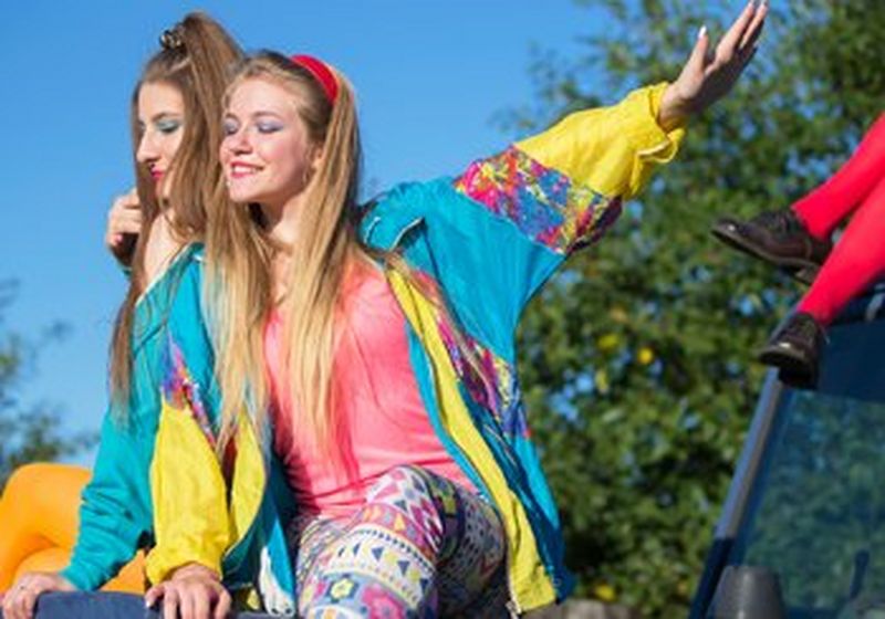 Two long-haired young women sit on the front of a jeep, smiling. They wear brightly coloured nineties-style shell suit jackets with bright leggings, one in bright acid orange and another in primary-coloured geometric shapes. They wear bright make up to match. The sky is bright clue and clear, and there are trees behind them. To their right, on the roof of the jeep is another person, but only their legs can be seen and are clad in bright red tights and black brogues.