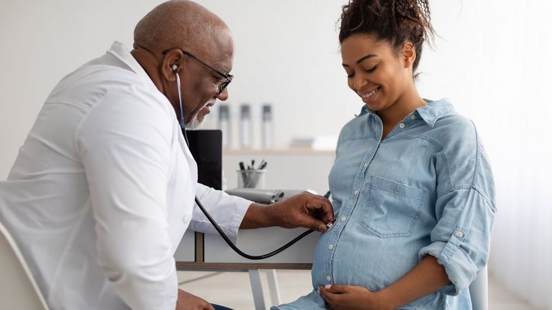 Un médecin en blouse blanche se penche vers l'avant pour examiner à l'aide de son stéthoscope le ventre d'une femme enceinte souriante, portant une chemise en jean.