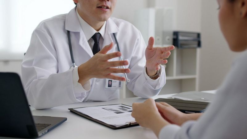A doctor in a white coat, with a stethoscope around his neck, gestures across a desk to a patient. The patient is blurred on the right-hand side of the picture. There is a laptop sat on the desk to the left of the picture.