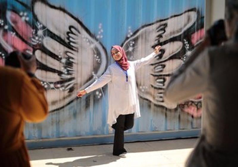 A young girl in a pink headscarf stands in front of graffiti of angel wings, so that they look like they are hers. On either side of her, with backs to the camera, are two people taking her photo.