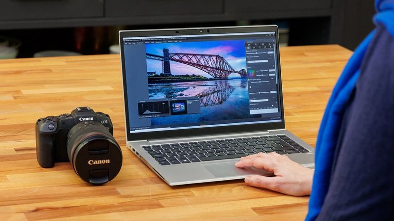 A woman editing a picture of a bridge at sunset in DPP on a laptop. A camera is next to the laptop on the table.