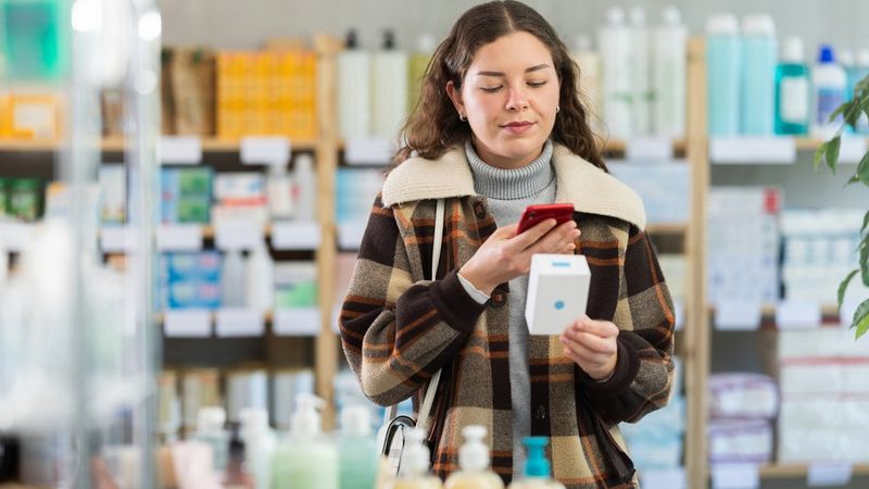 Een jonge vrouw houdt een product in een doos in één hand en scant het met haar smartphone. Planken met producten voor persoonlijke verzorging of apotheekproducten zijn zichtbaar op de achtergrond.