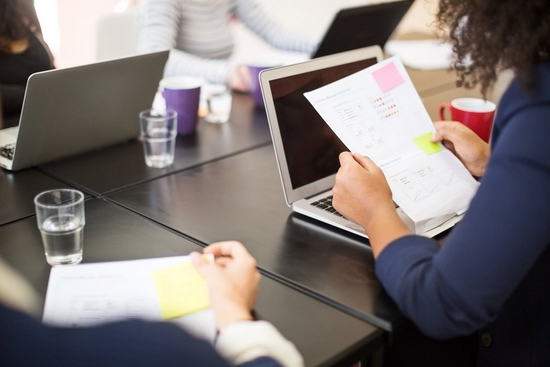 Three silver laptops on a black table with purple mugs and water. Woman reads a paper with a pink sticky note and red mug.