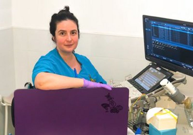 A dark haired woman in blue scrubs and purple latex gloves sits in front of a computer screen with assorted medical devices surrounding it. She has turned from the screen to pose for the camera.