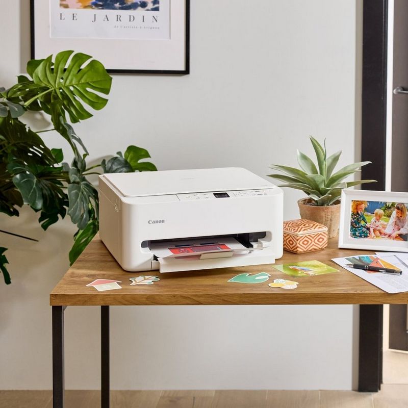 Canon-printer på et skrivebord i tre med fotoutskrifter, kunsthåndverksutstyr og en bordlampe på et kreativt arbeidsområde.
